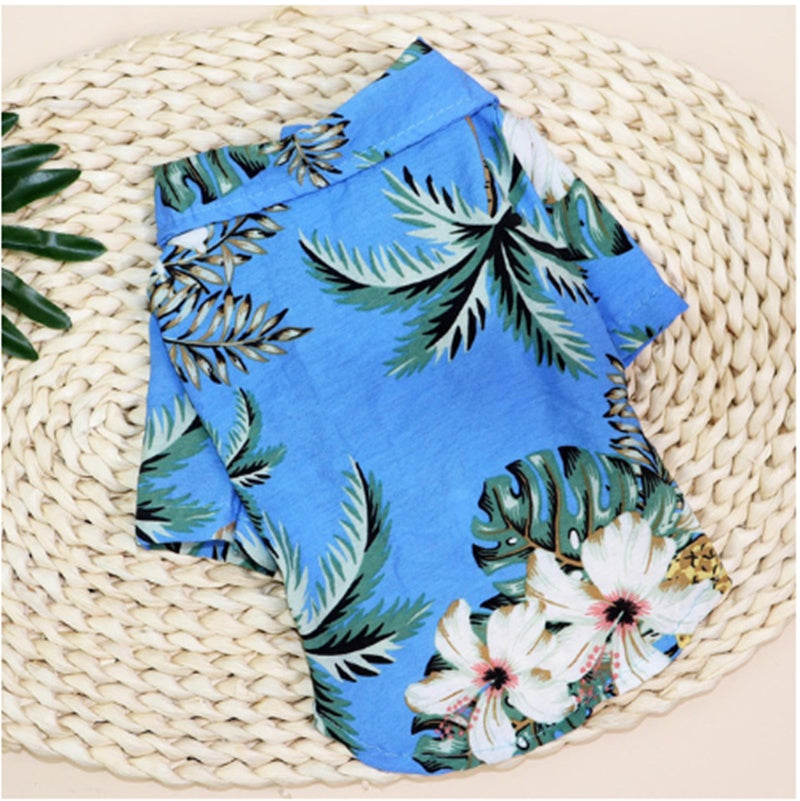 Folded blue tropical pet shirt with green palm leaves and white hibiscus flowers on a woven mat
