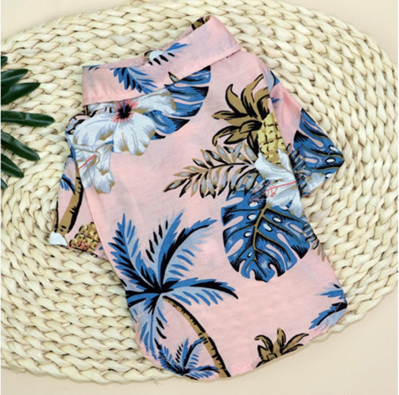 Pink tropical pet shirt with blue palm leaves and pineapple print displayed on a woven mat